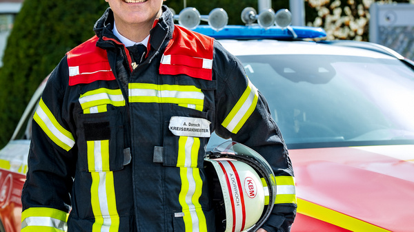 Ein Feuerwehrmann steht lächelnd vor einem Einsatzfahrzeug. Er trägt eine auffällige Feuerwehrjacke mit reflektierenden Streifen und hält einen Helm in der Hand. Im Hintergrund sind Bürobauten und grüne Bäume sichtbar.