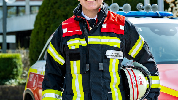 Ein Feuerwehrmann steht lächelnd vor einem Einsatzfahrzeug. Er trägt eine auffällige Feuerwehrjacke mit reflektierenden Streifen und hält einen Helm in der Hand. Im Hintergrund sind Bürobauten und grüne Bäume sichtbar.
