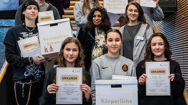 Eine Gruppe von zehn Personen steht auf einer Treppe und hält Zertifikate in den Händen. Die Themen der Zertifikate sind vertrauliche Beratung und körperliche Untersuchung. Im Hintergrund sind Holzgestelle und Fenster zu sehen. Die Stimmung ist feierlich und stolz.