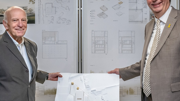 Zwei Männer stehen nebeneinander und präsentieren ein Architekturmodell auf einem Tisch. Im Hintergrund sind großformatige Pläne und Entwürfe sichtbar. Die Männer lächeln und scheinen stolz auf das Projekt zu sein.