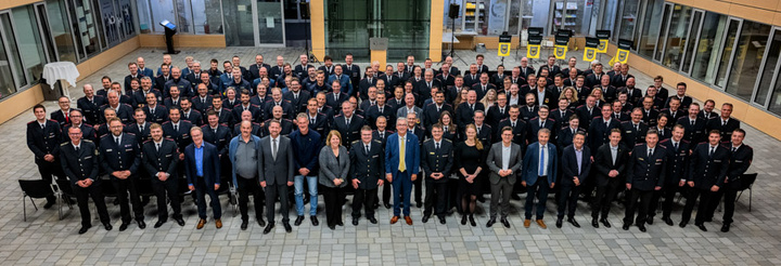 Gruppenfoto von über 100 Personen in uniformierten Kleidungen, die in einem hellen, gut beleuchteten Raum stehen. Die Menschen sind in mehreren Reihen angeordnet, einige lächeln, andere schauen ernst. Im Hintergrund sind Wände aus Glas und einige Stühle sichtbar.