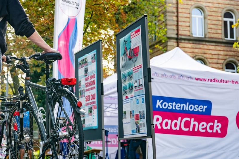 Eine Person steht neben einem Fahrrad auf einem herbstlichen Platz, umgeben von buntem Laub. Im Hintergrund sind Informationsstände mit Plakatwänden und ein Zelt, das für einen kostenlosen Radcheck wirbt. Die Atmosphäre ist freundlich und einladend. (Text KI generiert)