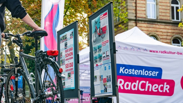 Eine Person steht neben einem Fahrrad auf einem herbstlichen Platz, umgeben von buntem Laub. Im Hintergrund sind Informationsstände mit Plakatwänden und ein Zelt, das für einen kostenlosen Radcheck wirbt. Die Atmosphäre ist freundlich und einladend. (Text KI generiert)