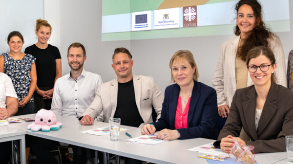 Eine Gruppe von zehn Personen sitzt und steht um einen Tisch. Im Hintergrund ist eine Präsentation mit dem Titel „Projekt Oktopus“ zu sehen. Die Teilnehmer scheinen an einem Arbeitsmeeting oder einer Präsentation teilzunehmen. Auf dem Tisch liegen verschiedene Unterlagen und ein Plüschtier.