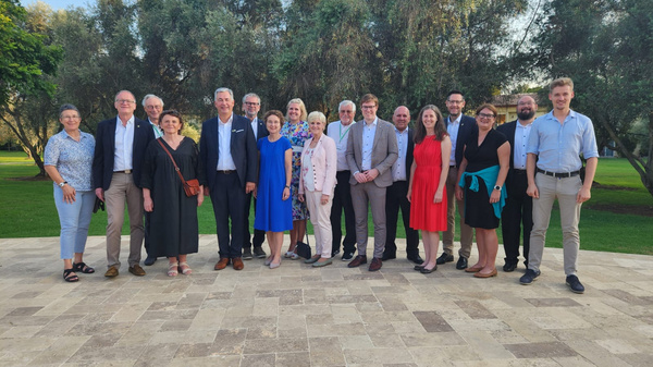 Eine Gruppe von 18 Personen steht auf einem eleganten Steinplatz im Freien, umgeben von Bäumen. Die Anwesenden tragen unterschiedliche, festliche Kleidung. Sie lächeln und posieren für ein Gruppenfoto, das eine fröhliche Atmosphäre vermittelt.