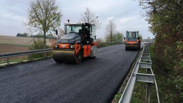 Two rollers are working on a freshly paved road, smoothing the asphalt. The scene includes trees along the roadside, with a rural landscape visible in the background. The sky is overcast, indicating possible rainy weather.