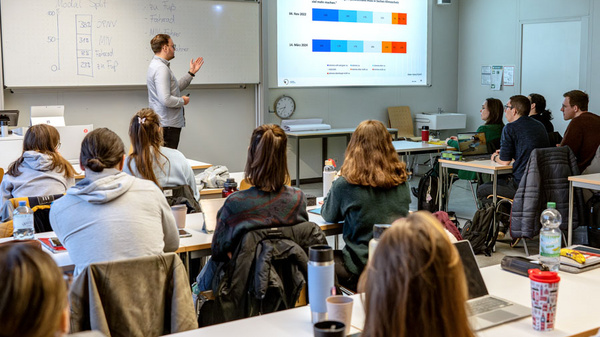 Ein Dozent präsentiert in einem Klassenzimmer vor einer Gruppe von Studierenden. Auf einer Leinwand sind Diagramme und Grafiken zu sehen, die Informationen deutlich hervorheben. Die Studierenden sind aufmerksam und auf ihre Laptops fokussiert.