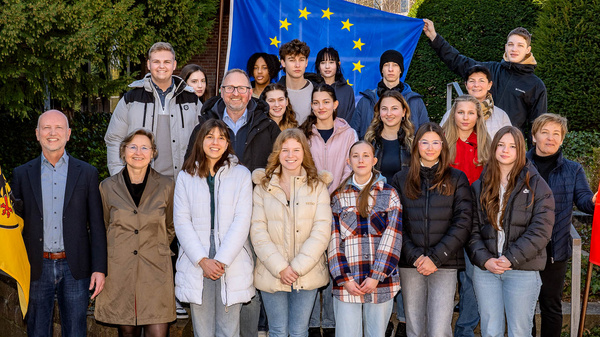 Eine Gruppe von etwa 20 Personen steht draußen vor grünen Hecken. Im Hintergrund ist eine europäische Flagge zu sehen. Links und rechts der Gruppe stehen Fahnen. Die Anwesenden lächeln und scheinen an einem gemeinsamen Projekt teilzunehmen.