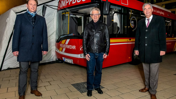 Drei Männer stehen vor einem roten Bus, der als Impfstation dient. Sie tragen formelle Kleidung und lächeln. Der Bus ist mit der Aufschrift „IMPFBUS“ und „FEUERWEHR“ gekennzeichnet. Im Hintergrund ist ein Zelt zu sehen, das den Impfbus schützt. (Text KI generiert)