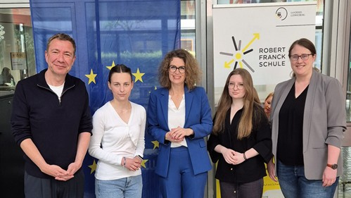 Eine Gruppe von fünf Personen steht lächelnd vor einer Europäischen Union-Flagge und einem Banner der Robert-Schuman-Schule. Sie tragen formelle Kleidung und scheinen an einer Veranstaltung oder Diskussion teilgenommen zu haben. Im Vordergrund sind ein Tisch mit Wassergläsern und einem Mikrofon zu sehen.