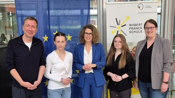 Eine Gruppe von fünf Personen steht lächelnd vor einer Europäischen Union-Flagge und einem Banner der Robert-Schuman-Schule. Sie tragen formelle Kleidung und scheinen an einer Veranstaltung oder Diskussion teilgenommen zu haben. Im Vordergrund sind ein Tisch mit Wassergläsern und einem Mikrofon zu sehen.