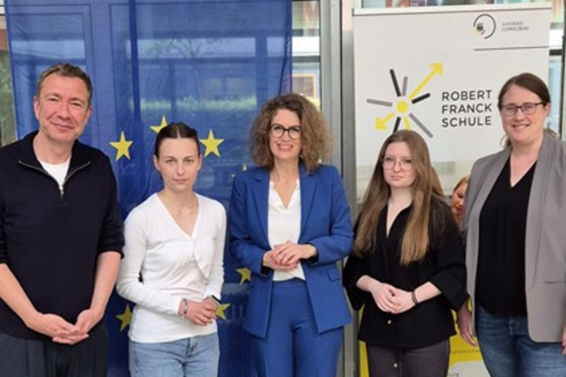 Eine Gruppe von fünf Personen steht lächelnd vor einer Europäischen Union-Flagge und einem Banner der Robert-Schuman-Schule. Sie tragen formelle Kleidung und scheinen an einer Veranstaltung oder Diskussion teilgenommen zu haben. Im Vordergrund sind ein Tisch mit Wassergläsern und einem Mikrofon zu sehen.