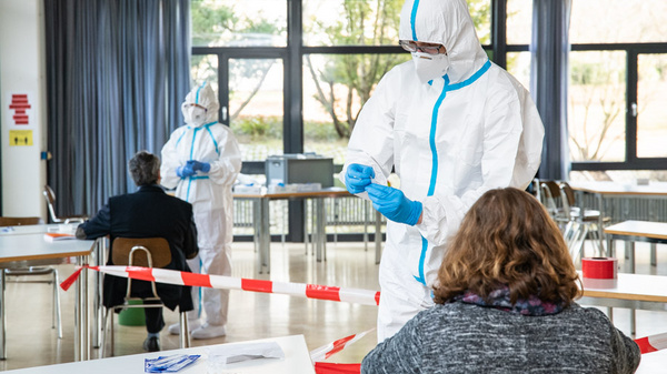 Two medical personnel in protective suits assist individuals in a room set up for testing. One person sits with their back to the viewer, while another staff member prepares equipment. The environment is organized, with safety measures in place.