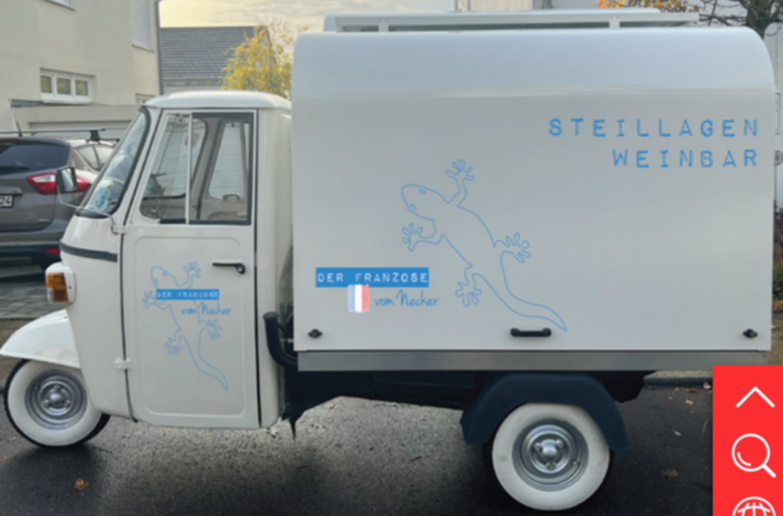 Ein dreirädriges Fahrzeug in weiß, das als Weinbar dient. Die Beschriftung zeigt den Namen "Der Franzose von Nacher" und enthält ein stilisiertes Bild eines Geckos. Auf der Seite steht "Steillagen Weinbar". Das Fahrzeug steht an einer Straßenecke und sieht einladend aus.