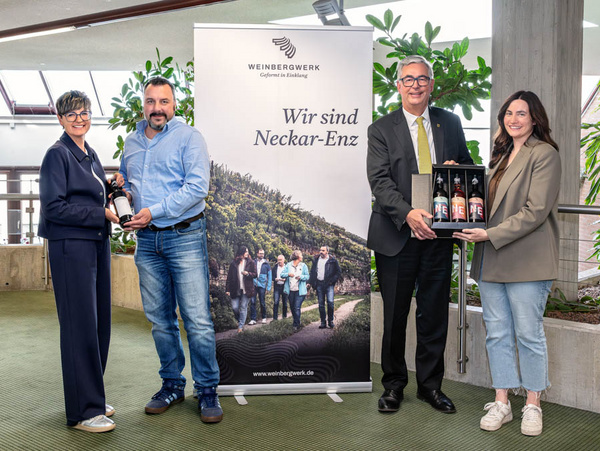 27.04.2026 - Der 1.Steillagenwein des Weinbergwerks wird übergeben Das Bild zeigt vier Personen, die in einem modernen Raum stehen. Zwei halten eine Flasche Wein, während die anderen eine Geschenkbox mit Flaschen präsentieren. Im Hintergrund steht ein Banner mit dem Schriftzug „Wir sind Neckar-Enz“ und einem Bild von einer Weinberglandschaft.