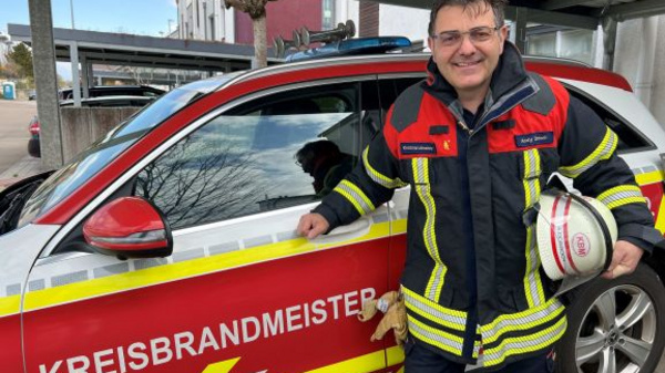 Ein Feuerwehrmann in Einsatzkleidung steht lächelnd neben einem Einsatzfahrzeug mit der Aufschrift „KREISBRANDMEISTER“. Er hält einen Feuerwehrhelm in der Hand. Im Hintergrund ist eine städtische Umgebung zu sehen. Das Bild vermittelt ein Gefühl von Einsatzbereitschaft und Teamgeist.