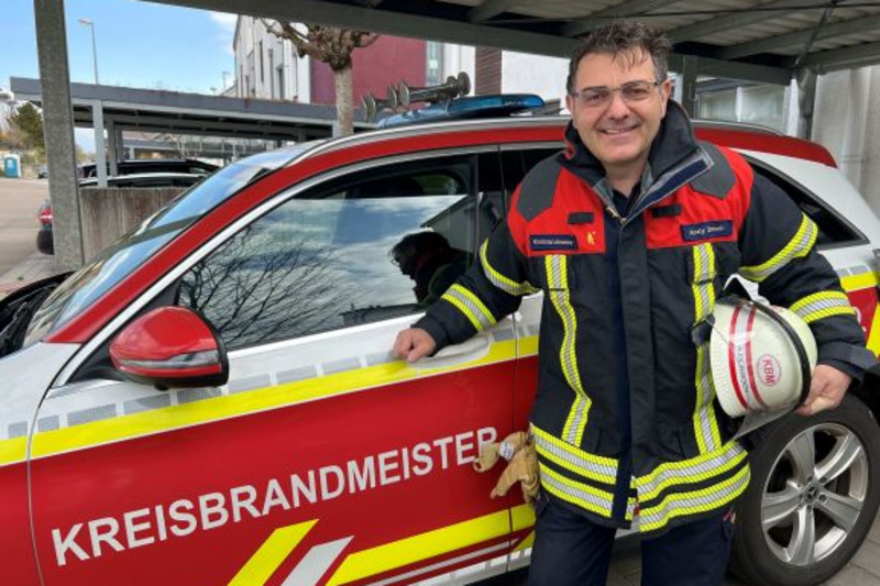 Ein Feuerwehrmann in Einsatzkleidung steht lächelnd neben einem Einsatzfahrzeug mit der Aufschrift „KREISBRANDMEISTER“. Er hält einen Feuerwehrhelm in der Hand. Im Hintergrund ist eine städtische Umgebung zu sehen. Das Bild vermittelt ein Gefühl von Einsatzbereitschaft und Teamgeist.