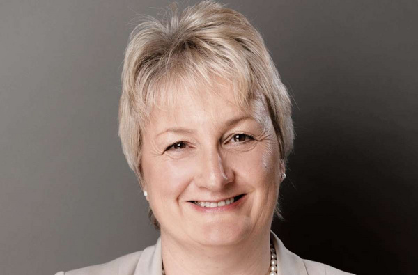 Porträt einer lächelnden Frau mit kurzem, blonden Haar. Sie trägt eine helle Bluse und eine Perlenkette. Der Hintergrund ist grau. Das Bild vermittelt eine freundliche und professionelle Ausstrahlung.