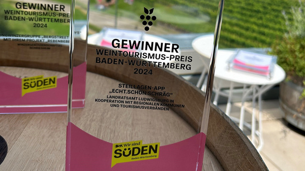 Ein transparenter Preis mit der Aufschrift "Gewinner Weintourismus-Preis Baden-Württemberg 2024" steht auf einem Holztisch. Im Hintergrund sind Weinreben und eine grüne Landschaft sichtbar. Der Preis gehört zur Kategorie "Steigenberger-App - Lofts in Constance".