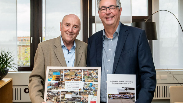 Zwei Männer stehen lächelnd in einem Büro. Der linke Mann hält ein gerahmtes Bild mit mehreren Fotos, während der rechte Mann ein Dokument in der Hand hält. Beide tragen formelle Kleidung und scheinen eine besondere Auszeichnung oder Feier abzuhalten. Hintergrund zeigt große Fenster und moderne Büromöbel.
