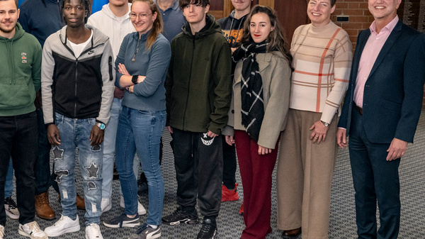 Eine Gruppe von 16 Personen steht in einem Raum zusammen, unterstützt von verschiedenen Flaggen im Hintergrund. Die Anwesenden sind unterschiedlich gekleidet und lächeln in die Kamera. Die Atmosphäre wirkt freundlich und informell, möglicherweise handelt es sich um ein Treffen oder eine Veranstaltung.