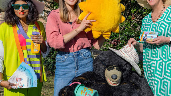 Drei Frauen stehen im Freien und lächeln. Eine Frau hält eine gelbe Sonne, während die andere ein Plüschtierbär mit Hut präsentiert. Die Frauen tragen Sonnenhüte und gestalten einen fröhlichen, sommerlichen Eindruck. Sie haben Flyer oder Broschüren in der Hand.