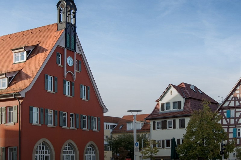 Das Bild zeigt eine Stadtansicht von Asperg mit einem historischen Gebäude auf der linken Seite, das eine markante Turmspitze hat. Im Hintergrund sind weitere traditionelle Häuser und Bäume zu sehen. Ein Text in der unteren rechten Ecke fordert dazu auf, die Region zu entdecken. (Text KI generiert)