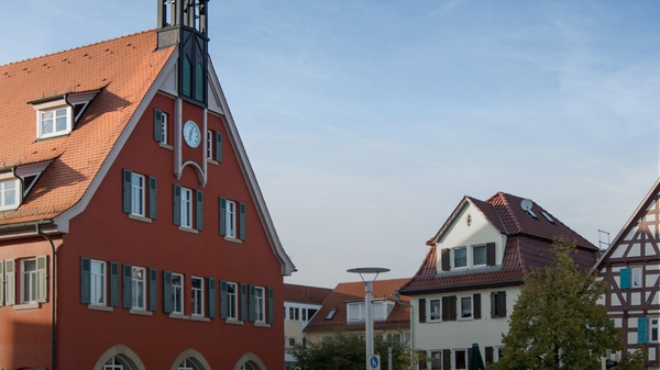 Das Bild zeigt eine Stadtansicht von Asperg mit einem historischen Gebäude auf der linken Seite, das eine markante Turmspitze hat. Im Hintergrund sind weitere traditionelle Häuser und Bäume zu sehen. Ein Text in der unteren rechten Ecke fordert dazu auf, die Region zu entdecken. (Text KI generiert)