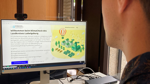 Ein junger Mann sitzt vor einem Computerbildschirm, auf dem eine Webseite mit dem Titel "Willkommen beim KlimaCheck des Landkreises Ludwigsburg" angezeigt wird. Neben Text sind grafische Elemente zu sehen, die mit Umwelt und Klimaschutz in Verbindung stehen.