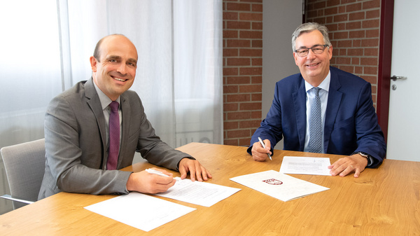 Deux hommes souriants, assis à une table en bois, signent des documents. L'un porte un costume gris avec une cravate violette, l'autre un costume bleu. Ils semblent engagés dans une conversation professionnelle, entourés d'un environnement de bureau simple.