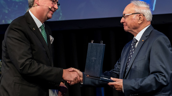 Two men are standing on stage, shaking hands. One man in a dark suit presents a blue award or plaque to the other man, who is wearing a light blue suit and glasses. The background appears to be a formal event setting with lights.