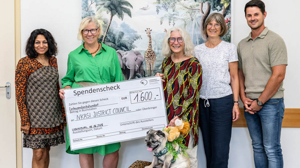 Eine Gruppe von fünf Personen steht zusammen, darunter eine Frau mit einem Scheck über 1.600 Euro für den Nkasi District Council. Neben ihnen sitzt ein Hund. Im Hintergrund sind afrikanische Tierbilder zu sehen.