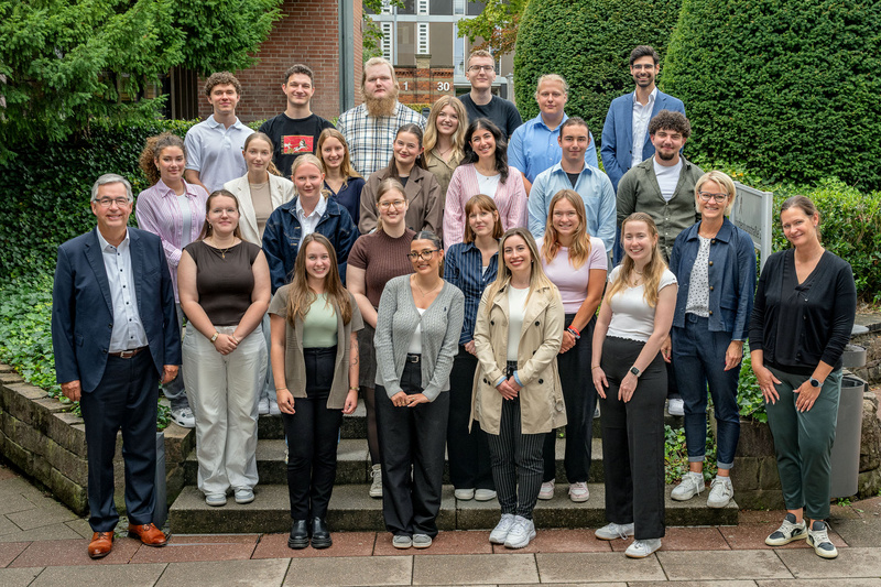 Eine Gruppe von etwa 30 Personen steht auf einer Treppe in einem Garten. Sie sind in professioneller Kleidung. Im Vordergrund steht ein älterer Mann mit Brille und zwei Frauen in hellem und dunklem Outfit. Die Atmosphäre ist freundlich und gemeinschaftlich.