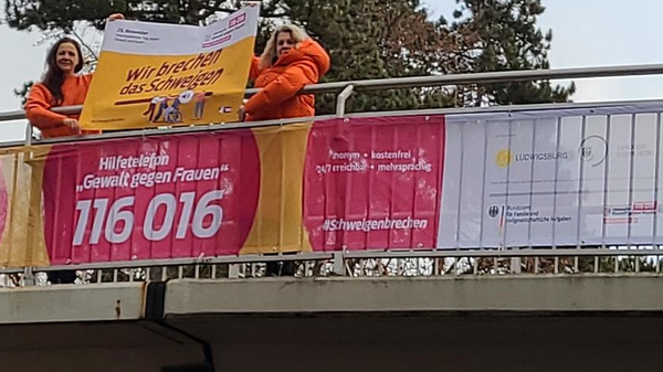 Zwei Personen in orangefarbener Kleidung stehen auf einer Brücke und halten ein großes Banner mit der Aufschrift „Wir unterstützen das Schutztelefon“. Zusätzlich sind Informationen zu einem Hilfsangebot für Gewaltopfer sowie eine Kontaktnummer sichtbar.