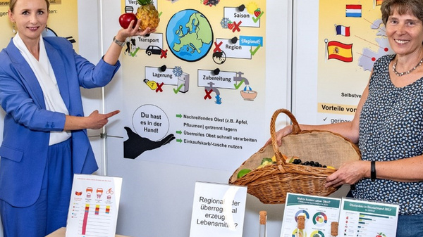 Due donne si trovano di fronte a pannelli informativi su acquisti di frutta e sostenibilità. Una donna tiene una mela, mentre l'altra indica una mappa. Su un tavolo ci sono frutta e documenti. I pannelli trattano di tematiche come l'origine del cibo e il suo impatto climatico.