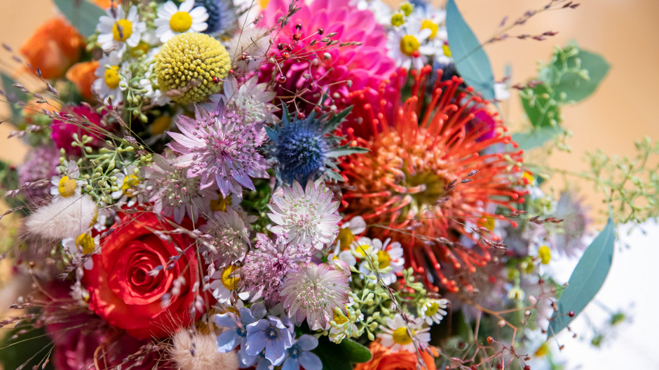A vibrant bouquet of mixed flowers, including roses, eucalyptus, and various wildflowers. The arrangement features a mix of colors such as red, pink, purple, yellow, and blue, creating a lively and cheerful display.