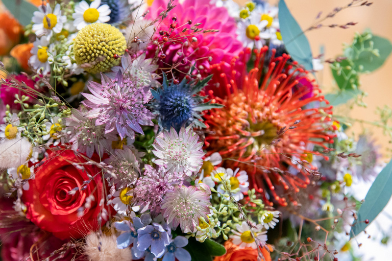 A vibrant bouquet of mixed flowers, including roses, eucalyptus, and various wildflowers. The arrangement features a mix of colors such as red, pink, purple, yellow, and blue, creating a lively and cheerful display.
