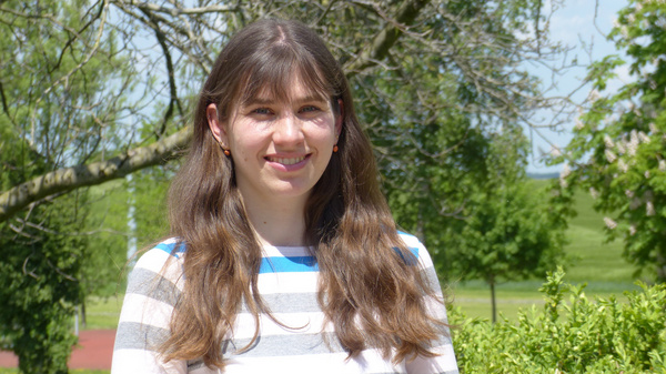 A young woman with long brown hair is smiling, standing outdoors in a park. She is wearing a striped shirt and there are trees and green grass in the background, suggesting a bright, sunny day.