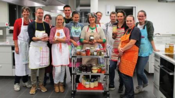 A group of twelve people, smiling and wearing aprons, gather around a cart filled with jars and canned goods in a modern kitchen. They appear to be celebrating a cooking or preservation activity, showcasing their teamwork and culinary creation.