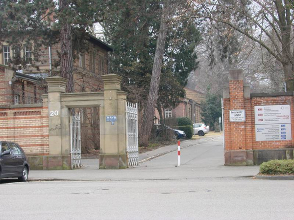 Ein Eingang zu einem Gelände, gekennzeichnet durch ein großes Tor. Auf der linken Seite sind Gebäude sichtbar, umgeben von Bäumen. Auf der rechten Seite steht ein Informationsschild mit verschiedenen Angaben und einem Parkverbotsschild. Es herrscht eine ruhige, städtische Atmosphäre.