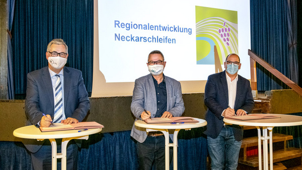 Three men in masks stand at white tables, signing documents. A presentation screen in the background displays the text "Regionalentwicklung Neckarschleifen." The setting appears to be a formal event.