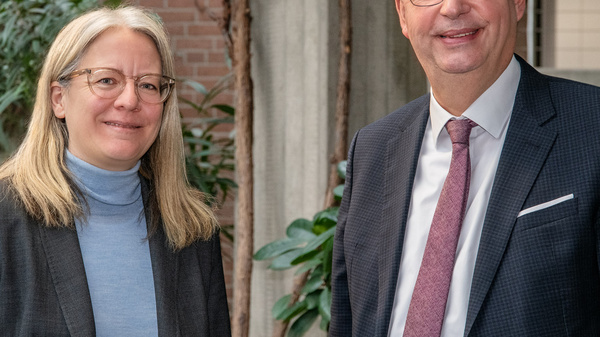Zwei Personen stehen lächelnd nebeneinander in einem Raum mit Pflanzen im Hintergrund. Die Frau hat lange blonde Haare und trägt einen blauen Pullover sowie einen grauen Blazer. Der Mann trägt ein dunkles Sakko mit einer Krawatte und wirkt freundlich.