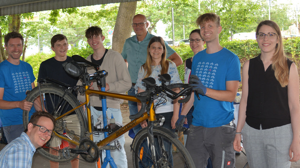 Un gruppo di otto persone sorride vicino a una bicicletta in riparazione. Alcuni indossano magliette blu e altri vestiti casual. L'ambiente è all'aperto, sotto una struttura con alberi sullo sfondo. L'atmosfera è di convivialità e collaborazione.