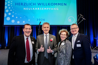 Vier Personen stehen lächelnd vor einer Bühne mit einem Banner, das "Herzlich Willkommen Neujahrsempfang 2026" zeigt. Die Personen tragen formelle Kleidung und präsentieren positive Gesten, während im Hintergrund ein Mikrofon steht.
