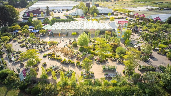 Die Luftaufnahme zeigt einen Garten mit vielen Pflanzen, Bäumen und Blumenbeeten. Im Hintergrund sind Gewächshäuser und Lagerhallen zu sehen. Der Garten ist ordentlich angelegt und lädt zum Spaziergang ein. Die Umgebung wirkt ruhig und grün, ideal für Gartenliebhaber.