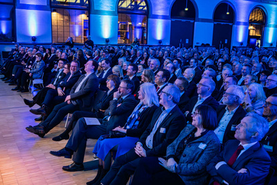 Eine große Menschenmenge sitzt in einem beleuchteten Raum, die meisten Personen lachen oder lächeln. Die Atmosphäre wirkt festlich und fröhlich. Die Teilnehmer scheinen an einer Veranstaltung teilzunehmen, möglicherweise einer Rede oder Präsentation.