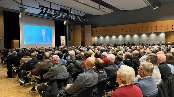 Eine große Menschenmenge sitzt in einem Veranstaltungssaal. Auf der Bühne ist ein Redner zu sehen, der auf einen Bildschirm schaut, auf dem sein Bild projiziert wird. Die Zuhörer wirken aufmerksam und konzentriert auf die Präsentation.