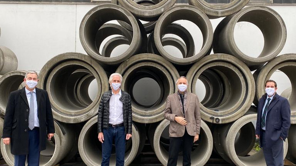 Four individuals wearing masks stand in front of a stack of large concrete pipes. The setting appears to be an industrial area, with the pipes arranged in a neat formation behind them. They are dressed in formal and casual attire, and the mood is professional.