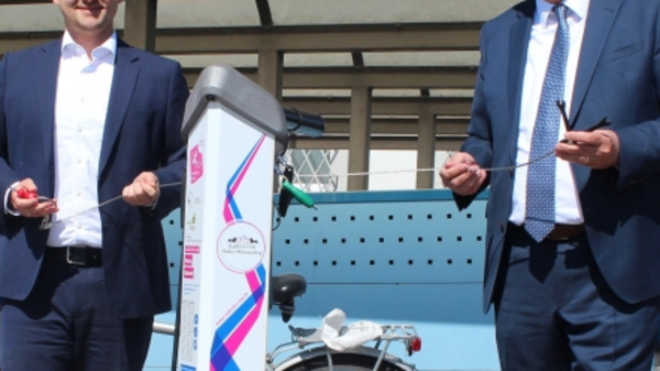 Two men in business attire stand beside a bike-sharing station. They are smiling and holding tools, suggesting an inauguration or launch event. A bicycle is parked in the background, and the station has multiple bike slots. The setting appears to be outdoor, likely in an urban area.