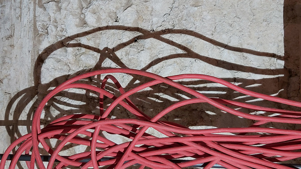A collection of tangled pink cables lies on a textured surface, casting intricate shadows on the wall behind them. The interplay of light emphasizes the curves and bends of the cables, creating a visually striking contrast between the cables and the wall.